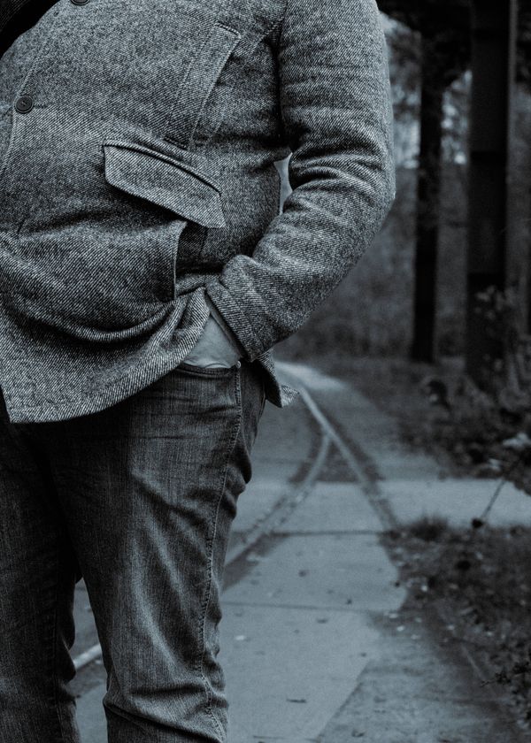 Person standing. Filling half the frame. One hand in trouser pocket. Wool trench coat. Jeans. Other half of frame is blurry. It shows old tram lines and trees.